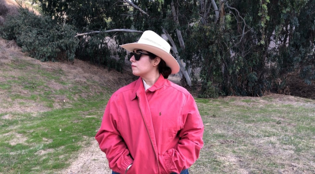 Image of Flores in nature. They are wearing a cowboy hat, sunglasses, and a jacket. 